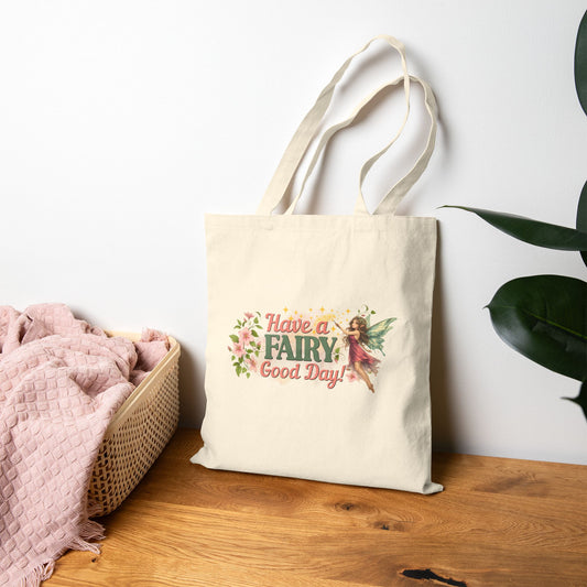 A cream canvas tote bag with a colorful 'Have a FAIRY Good Day!' graphic, standing on a wooden table next to a pink blanket and a plant against a white wall.