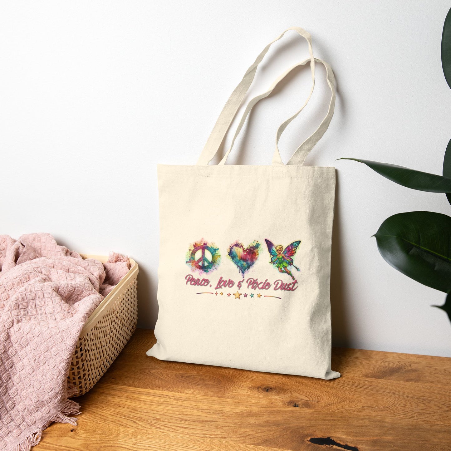 A cream canvas tote bag with colorful peace-themed graphics and text, resting on a wooden table next to a pink blanket and a plant against a white wall.