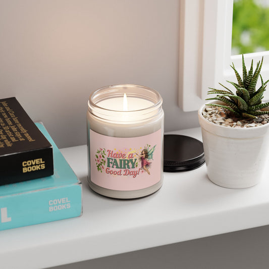 A lit candle in a clear glass jar sits on a white windowsill next to a small potted succulent and stacked books. The candle has a pink label that reads 'Have a FAIRY Good Day!' with a fairy illustration.
