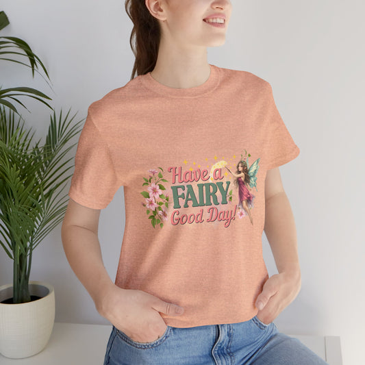 A woman wearing a heathered peach t-shirt with a colorful graphic that reads 'Have a FAIRY Good Day!' accompanied by a fairy and floral illustrations, paired with blue jeans in a bright indoor setting with a potted plant.